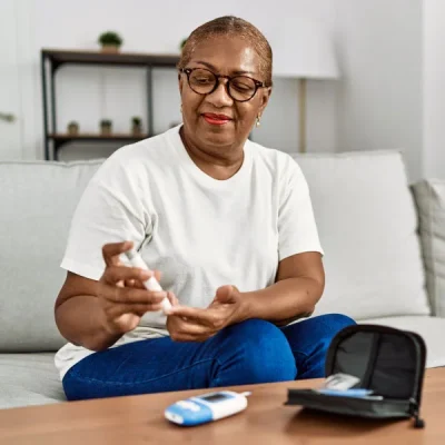 Older woman wearing glasses sits on a sofa at home, using a glucometer to test blood sugar with a test strip nearby.