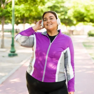 Woman in a purple athletic jacket wearing white headphones, smiling while standing in a sunny park.